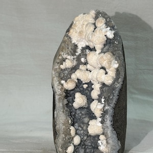 Könnte beinhalten: Ein vertikales Geoden-Exemplar mit einem dunkelgrauen, kristallinen Innenraum, der mit kleinen, scharfen Spitzen ausgekleidet ist. Die Mitte ist mit Clustern aus abgerundeten, cremefarbenen Mineralformationen verziert, die sich vom dunklen, strukturierten Stein abheben.