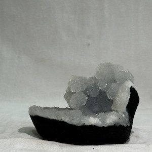 Op de afbeelding: Een halvemaanvormig geode-exemplaar met een donkere, matzwarte buitenkant en een contrasterend interieur gevuld met clusters van doorschijnende, sprankelende witte kwartskristallen die het licht vangen op het onregelmatige oppervlak.