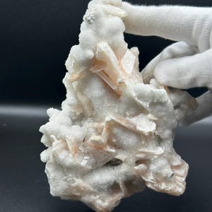 Könnte beinhalten: Ein Mineralexemplar mit einer Ansammlung weißer, kristalliner Strukturen und eingebetteten, blass pfirsichfarbenen, klingenartigen Formationen. Die Oberfläche ist mit einer feinen, funkelnden, körnigen Textur bedeckt, die ein gezacktes Aussehen erzeugt.
