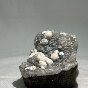 Peut inclure: Un spécimen minéral composé d'une base sombre et rugueuse surmontée d'un amas de petits cristaux gris translucides. Parmi ces cristaux gris se trouvent plusieurs formations blanches opaques et arrondies à l'aspect sphérique texturé.