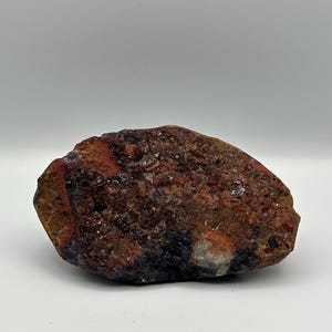 Könnte beinhalten: Ein raues, unregelmäßiges Mineralexemplar mit einer texturierten Oberfläche aus dunkelroten, braunen und schwarzen kristallinen Formationen. Der Stein zeigt eine Mischung aus erdigen Tönen und kleinen, reflektierenden Mineralablagerungen.