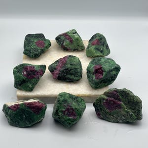 Op de afbeelding: Een verzameling van acht ruwe robijn in zoisiet stenen, elk met een unieke combinatie van diep magenta-rode robijnkristallen ingebed in een textuurrijke, gevlekte groene en zwarte zoisiet matrix, gerangschikt in een groep op een vierkante basis.