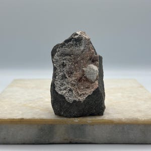 Peut inclure: Un spécimen de roche volcanique gris foncé présentant une cavité centrale remplie d'un dépôt minéral rose clair texturé et un petit amas de cristaux blancs sphériques faisant saillie au centre de la surface rugueuse et irrégulière.