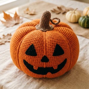 Op de afbeelding: Een ronde, oranje gehaakte pompoen met een bruine steel en een zwart jack-o'-lantern gezicht met twee driehoekige ogen, een kleine driehoekige neus en een brede, gebogen mond met twee kleine tandjes, allemaal gemaakt van garen met stiksels.