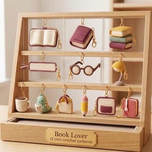 Puede incluir: Un expositor de madera clara con doce llaveros de ganchillo en miniatura con formas de libros, gafas, lámpara, taza, criatura, bolso y lápiz. Una placa dorada en la base muestra el texto Book Lover 12 mini crochet patterns en negro.