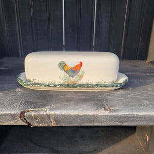 May include: A ceramic butter dish with a rooster design. The dish is rectangular with a lid and a matching base. The rooster is depicted in vibrant colors, with a red comb and a mix of blue, orange, and green feathers. The base has a green trim.