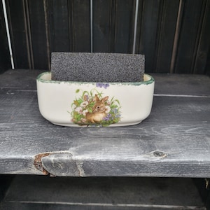 May include: White ceramic sponge holder with a green rim and a floral design featuring a brown rabbit. The holder is sitting on a dark gray wooden surface.