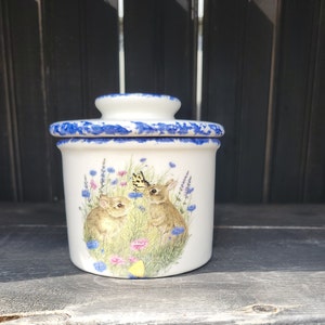May include: White ceramic butter dish with a blue rim and a floral design featuring two bunnies and a butterfly.