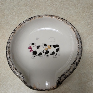 May include: White ceramic spoon rest with a distressed brown rim. The spoon rest features a whimsical illustration of two cows with black spots and bows. A charming kitchen accessory.