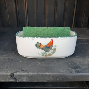 May include: White ceramic dish with a green rim and a rooster design. A green sponge is sitting inside the dish.