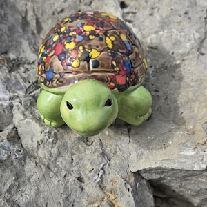 May include: A ceramic turtle figurine with a green head and feet, and a shell decorated with colorful spots. The turtle is set against a gray rock background, showcasing its whimsical design.