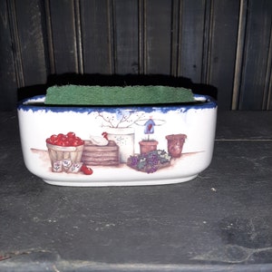 May include: White ceramic sponge holder with a blue rim and a country-themed design featuring a basket of red cherries, a white chicken, a potted plant, and a grapevine.