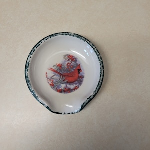 May include: A ceramic spoon rest with a green rim and a white background. The center features a red cardinal bird perched on a branch with red berries.