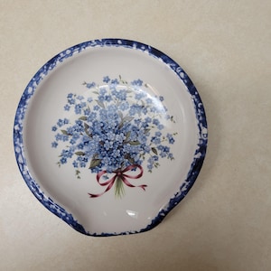 May include: A white ceramic spoon rest with a blue and white speckled border. The center features a bouquet of blue forget-me-not flowers with a red ribbon.