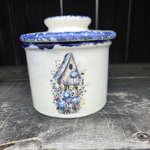 May include: A white ceramic jar with a lid, featuring a blue and white floral design. The jar has a birdhouse illustration with blue and yellow flowers. The lid and rim are decorated with a blue pattern. The jar is sitting on a wooden surface.