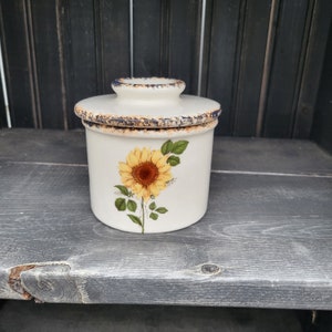 May include: A white ceramic butter dish with a lid, featuring a yellow sunflower design with green leaves. The rim and lid edge have a distressed blue and brown trim. The dish is on a gray wooden surface.