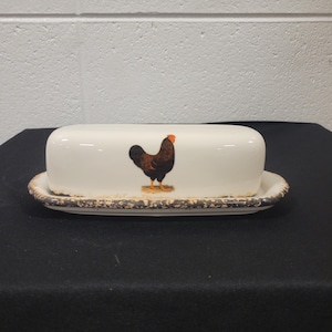 May include: White ceramic butter dish with a rooster design. The dish has a brown rooster with a red comb and wattle. The dish has a brown rim with a distressed look.