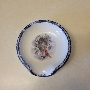 May include: White ceramic spoon rest with a blue rim. The center features a winter scene with a cat looking out a window, a red cardinal, and other birds. The spoon rest has a curved shape with a small lip.
