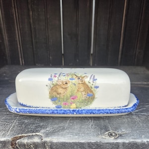 May include: A white ceramic butter dish with a blue trim and a design of two bunnies in a field of flowers.