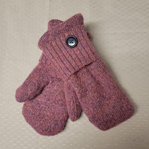 May include: A pair of pink and purple knit mittens with a button closure. The mittens are made of a textured yarn and have a ribbed cuff. The button is dark blue. The mittens are laying on a white surface.