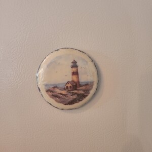 May include: A round, white magnet with a painted image of a lighthouse on a sandy beach with a small house in front of it. The lighthouse is red and white with a blue roof. There are birds flying in the sky.