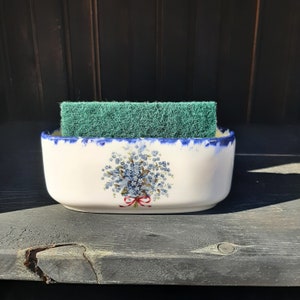 May include: A white ceramic sponge holder with a blue floral design and a red bow. The holder has a blue trim and contains a green scrub sponge. The holder is rectangular and sits on a dark wooden surface.