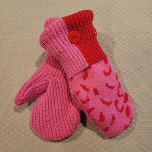 May include: A pair of knitted mittens in shades of pink and red. One mitten is solid pink, while the other features a pattern of red shapes on a pink background. A red button accents the cuff.