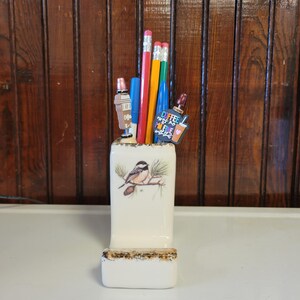 May include: A white ceramic pencil holder with a bird and pine branch design. The holder has a small shelf on the bottom. There are several pencils and pens in the holder, including a blue pen with the text "Coffee makes me feel alive" on it.