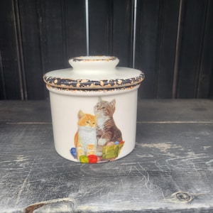 May include: A white ceramic butter dish with a lid, featuring a distressed brown rim. The dish is decorated with a colorful illustration of two cats and a ball of yarn. The lid has a small knob for easy lifting.