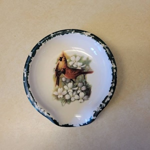 May include: A white ceramic spoon rest with a green rim. The spoon rest features a painted image of two red cardinals perched on a branch with white flowers.