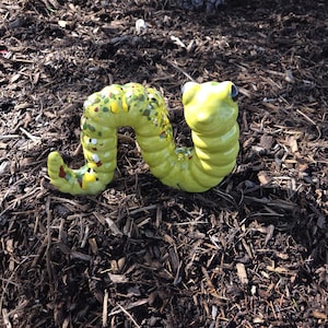 May include: A whimsical, ceramic worm sculpture in a bright, speckled yellow-green. The worm is posed in a curved shape, with a smiling face and black eyes. It rests on a bed of dark brown mulch, with a tree trunk in the background.