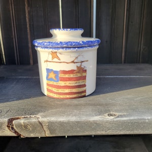 May include: A white ceramic crock with a blue lid and a painted American flag design. The flag has red, white, and blue stripes and stars. The text on the flag reads "HOLDS RED, WHITE, BLUE, BUT SO FRIENDLY WE HAILED ALL COLORS, BY THE RIVERS EARLY LIGHT, HOLD OUR HEAD WAS STILL THERE."
