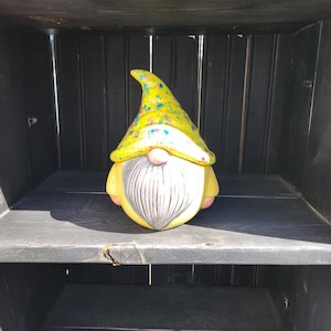 May include: A yellow ceramic gnome figurine with a colorful, speckled hat. The gnome has a white beard and a pink nose.
