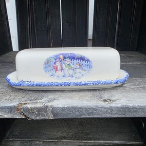 May include: White ceramic butter dish with a blue and white painted border. The lid features a winter scene with snowmen. The dish is rectangular with a rounded top and sits on a matching tray.