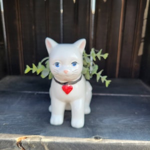 May include: White ceramic cat planter with blue eyes and a red heart on its chest. The planter is filled with green leafy plants.