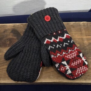 May include: A pair of dark gray knitted mittens with a red button detail. One mitten features a patterned design in red, black, and white. The mittens are displayed on a wooden surface, suggesting a cozy, winter theme.