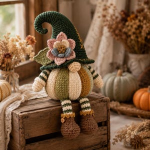Könnte beinhalten: Eine gehäkelte Gnomenfigur mit einer dunkelgrünen, spitzen Mütze, die mit einer großen rosa und blauen Blume verziert ist. Der Körper ist eine runde, gestreifte Kürbisform in Creme, Olivgrün und Braun, mit baumelnden, gestreiften Beinen und braunen Stiefeln.