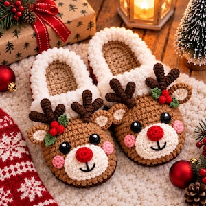 May include: A pair of brown crochet slippers featuring a reindeer face design with a bright red nose, black eyes, pink cheeks, and dark brown antlers. The slippers have a thick, white, textured cuff around the opening and are decorated with small green leaves and red berries.