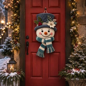 May include: A crocheted snowman door hanging featuring a blue top hat adorned with white flowers, green leaves, and red berries. The snowman has a round cream face, a small orange nose, pink cheeks, and a blue and white striped fringed scarf.