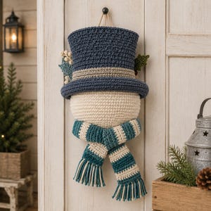 May include: A crocheted snowman wall hanging featuring a dark blue top hat with a light tan band, a cream-colored round head, and a teal and cream striped scarf with fringed ends. Decorative green leaves and small white berries accent the hat.