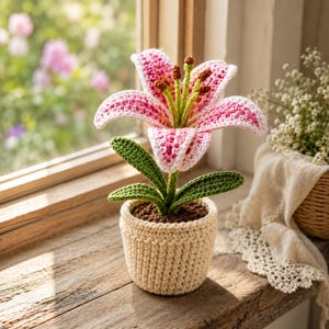 May include: A crocheted lily flower with pink and white petals, green stems, and brown stamens, potted in a textured beige yarn container. The plant features dark green leaves and is crafted with visible yarn stitches throughout the entire structure.