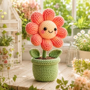 Könnte beinhalten: Eine gehäkelte Blume mit pfirsichfarbener Mitte, rosa Wangen und schwarzen Augen, umgeben von korallenrosa Blütenblättern. Die Blume hat einen grünen Stiel mit zwei Blättern und steht in einem strukturierten, hellgrünen Häkeltopf mit brauner Erde.