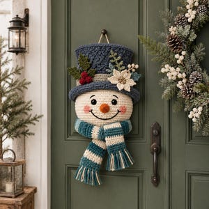 May include: A crocheted snowman wall hanging featuring a dark blue top hat adorned with green leaves, red berries, and a white flower. The snowman has a cream-colored face, a bright orange nose, and a blue and cream striped scarf with fringe.