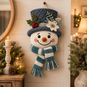May include: A crocheted snowman wall hanging featuring a cream-colored head, a dark blue top hat adorned with green leaves, red berries, and a white poinsettia, and a blue and white striped scarf with fringe, all crafted with textured yarn.
