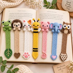 May include: Seven crocheted animal bookmarks featuring a green frog with a leaf, a brown hedgehog, a white cat with a paw print, a yellow bee, a pink pig with a curly tail, a blue elephant with a heart, and a brown owl with a feathered tail.