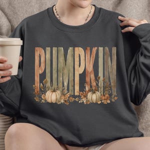 May include: A dark gray crewneck sweatshirt featuring the word PUMPKIN in large, vertical, wood-textured letters. The design includes watercolor-style illustrations of pumpkins, autumn leaves, and dried foliage at the base of the lettering.