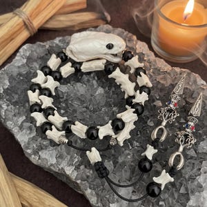 May include: A beaded bracelet featuring a white snake head focal piece, alternating white bone-shaped beads, and smooth black spherical beads. The bracelet includes two silver-toned metal charms with red and blue accents on black cords.