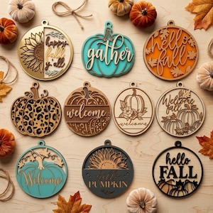 Può includere: Una collezione di undici ornamenti in legno circolari e a forma di zucca con temi autunnali. I disegni includono girasoli, zucche e motivi leopardati con testi come happy fall y'all, gather, hello fall, welcome e hello PUMPKIN.
