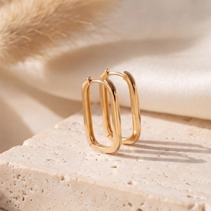 Könnte beinhalten: Ein Paar goldfarbene Creolen in rechteckiger, länglicher Form. Die Ohrringe haben eine glatte, polierte Metalloberfläche und einen Klappverschluss und stehen aufrecht auf einer strukturierten, hellen Steinoberfläche.