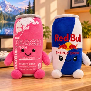 May include: Two plush toys shaped like Red Bull energy drink cans. The left toy is pink with white text reading The Peach Edition and White Peach. The right toy is blue and white with the Red Bull logo. Both have small arms, legs, and faces.